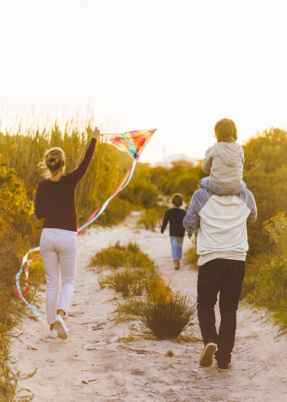 A photo of family walking on sandy footpath amidst plants. Father is carrying son on shoulders during summer. Rear view of woman flying kite in nature.